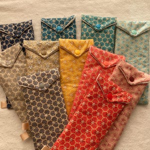 Peut inclure: Plusieurs pochettes en tissu faites à la main, de couleurs variées : bleu marine, sarcelle, or et rouge. Chaque pochette présente un motif géométrique et une fermeture à bouton. Les pochettes sont disposées sur une surface de couleur neutre.