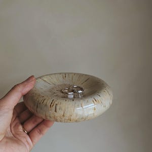 May include: A small, round ceramic dish with a speckled beige and brown glaze. Two silver rings rest inside the dish. The dish is held in a hand, showcasing its size and design. Ideal for holding jewelry or small items.