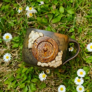 May include: A dark brown ceramic mug featuring a raised, circular wooden-textured door design in the center, surrounded by small, light-colored stone-like accents. The mug has a smooth handle and a mottled, earthy finish on its exterior surface.