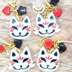 May include: Four white kitsune masks with red and black details, each attached to a gold charm with a crescent moon, a star, and a heart. The charms are connected to a white string.