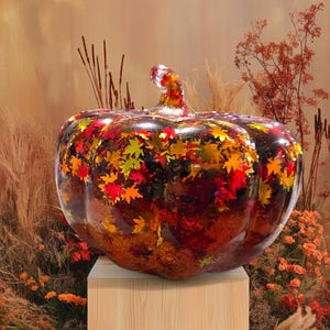 May include: A decorative pumpkin with a clear, glossy finish, filled with colorful leaf-shaped confetti. The pumpkin sits on a wooden pedestal, with a backdrop of autumnal foliage in shades of orange and brown.