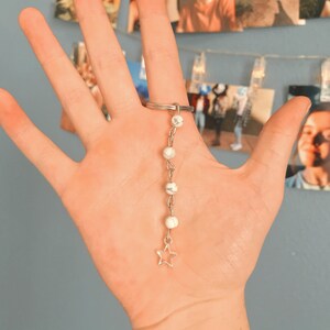 Può includere: Un anello d'argento con un ciondolo pendente. Il ciondolo presenta perline bianche e una piccola stella d'argento. L'anello è esposto su una mano, con un muro blu e foto sullo sfondo.