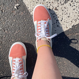 May include: Canvas sneakers with peach toe caps and heel panels, light blue side stripes, and white laces. The shoes are on a dark asphalt surface with a white crosswalk stripe.