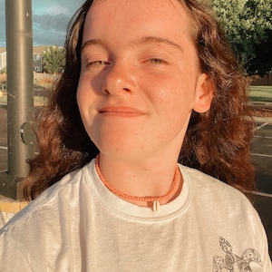 以下が含まれることがあります： 黒いウミガメのグラフィックが描かれた白いTシャツを着た、長い茶色の髪の若い人。ピンクのビーズネックレスに白い貝殻のペンダントをつけています。