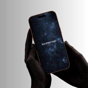 May include: A smartphone with a copper-colored frame and a screen displaying a dark blue galaxy background with white stars. The text "We are all made of stardust" is printed in white in the center of the screen. The device has a pill-shaped cutout.
