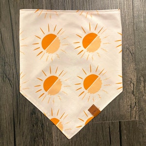 Puede incluir: Bandana blanca con un patrón repetido de soles naranjas con rayos amarillos. La bandana tiene una etiqueta de cuero marrón con el texto "The Wild" en ella.