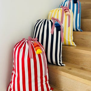 May include: Four striped fabric bags in red, navy, yellow, and blue, each with white stripes. The bags are tied with striped ribbons and adorned with colorful tassels. The bags are arranged on wooden stairs.