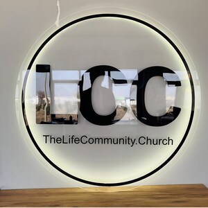 May include: A black and white circular sign with the letters "LCC" in a bold font. The text "TheLifeCommunity.Church" is written below the letters.