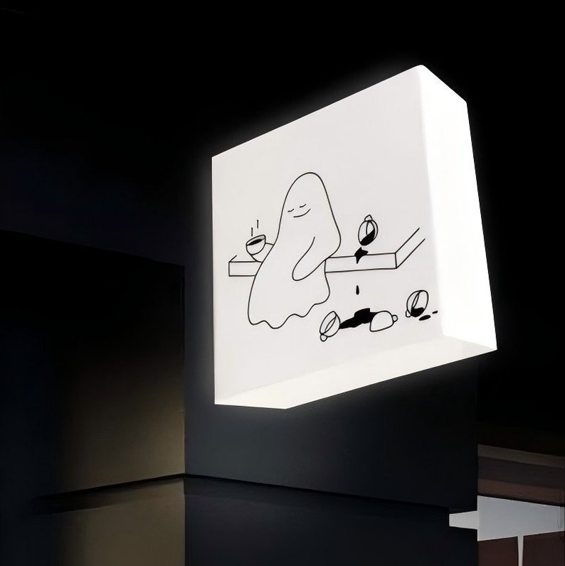 Op de afbeelding: Een witte, rechthoekige lamp met een zwart-wit illustratie van een spook dat aan een tafel zit met een kop koffie. Gemorste koffie en kopjes liggen op de tafel en de vloer. De lamp is aan een donkere muur bevestigd.