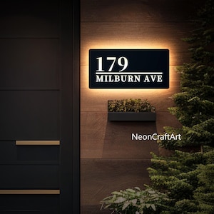 May include: A black and white illuminated house number sign with the number 179 and the street name Milburn Ave. The sign is mounted on a wooden wall with a small potted plant below it.