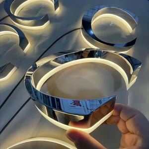 K&ouml;nnte beinhalten: Nahaufnahme eines silbernen Metallbuchstabens C mit einem wei&szlig;en LED-Lichtstreifen am Rand. Der Buchstabe ist beleuchtet und scheint Teil eines gr&ouml;&szlig;eren Schildes zu sein.