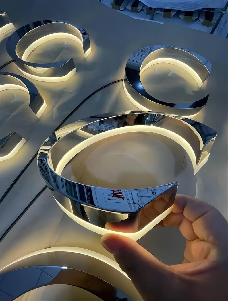 K&ouml;nnte beinhalten: Nahaufnahme eines silbernen Metallbuchstabens C mit einem wei&szlig;en LED-Lichtstreifen am Rand. Der Buchstabe ist beleuchtet und scheint Teil eines gr&ouml;&szlig;eren Schildes zu sein.