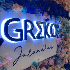 K&ouml;nnte beinhalten: Ein wei&szlig;es Schild mit schwarzer Schrift, auf dem "GREKO Jalandhar" steht, umgeben von blauen, rosa und wei&szlig;en Blumen.