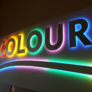 K&ouml;nnte beinhalten: Ein wei&szlig;es Schild mit dem Wort "COLOUR" in gro&szlig;en, erhabenen Buchstaben. Jeder Buchstabe ist in einer anderen Farbe des Regenbogens umrandet, beginnend mit Rot und endend mit Blau.