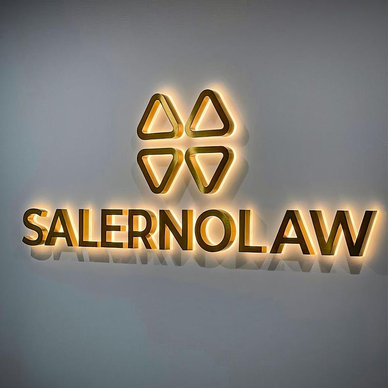 K&ouml;nnte beinhalten: Ein goldfarbenes Schild mit dem Text "SALERNOLAW" in einer fetten, serifenlosen Schrift. Das Schild zeigt auch ein stilisiertes Logo aus vier goldenen Dreiecken.