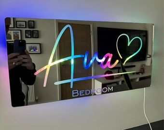 May include: A mirrored sign with a rainbow-colored inscription of "Ava" with a heart symbol and the word "BEDROOM" below. The sign is illuminated with colorful LED lights.