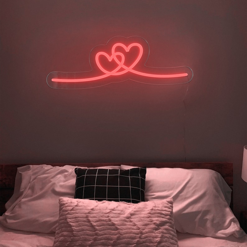 Op de afbeelding: Een neonbord met twee in elkaar verweven harten in een rode gloed, gemonteerd aan een muur boven een bed met witte beddengoed en kussens.