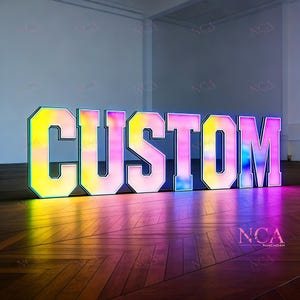 Puede incluir: Un letrero grande e iluminado que deletrea la palabra "CUSTOM" en letras de bloque en negrita. Las letras tienen un efecto degradado, que va del amarillo al rosa y al morado. El letrero está delineado en verde neón y se encuentra sobre un piso de madera.