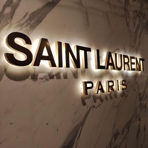 K&ouml;nnte beinhalten: Ein goldfarbenes Schild mit dem Text "SAINT LAURENT PARIS" an einer wei&szlig;en Marmorwand.