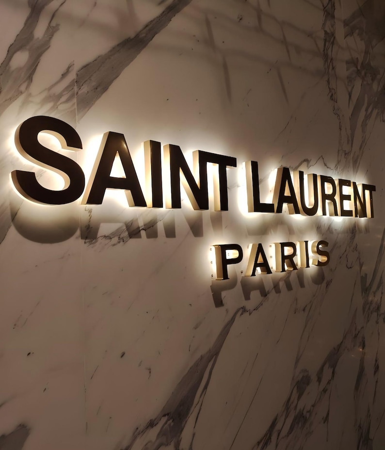 K&ouml;nnte beinhalten: Ein goldfarbenes Schild mit dem Text "SAINT LAURENT PARIS" an einer wei&szlig;en Marmorwand.