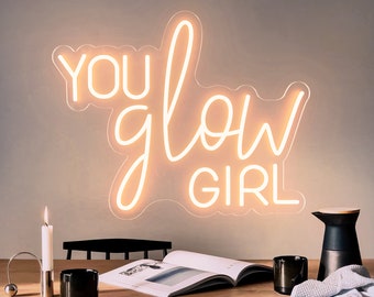 May include: White neon sign with the text "YOU glow GIRL" on a white wall. The sign is hanging above a wooden table with a book, candles, and mugs.