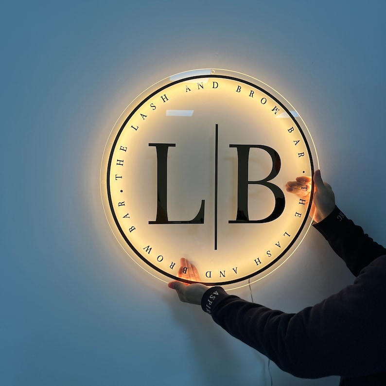 May include: A white circular sign with black text that reads "Lash and Brow Bar. The Lash and Brow Bar." The sign has a lit-up background with the letters "LB" in the center.