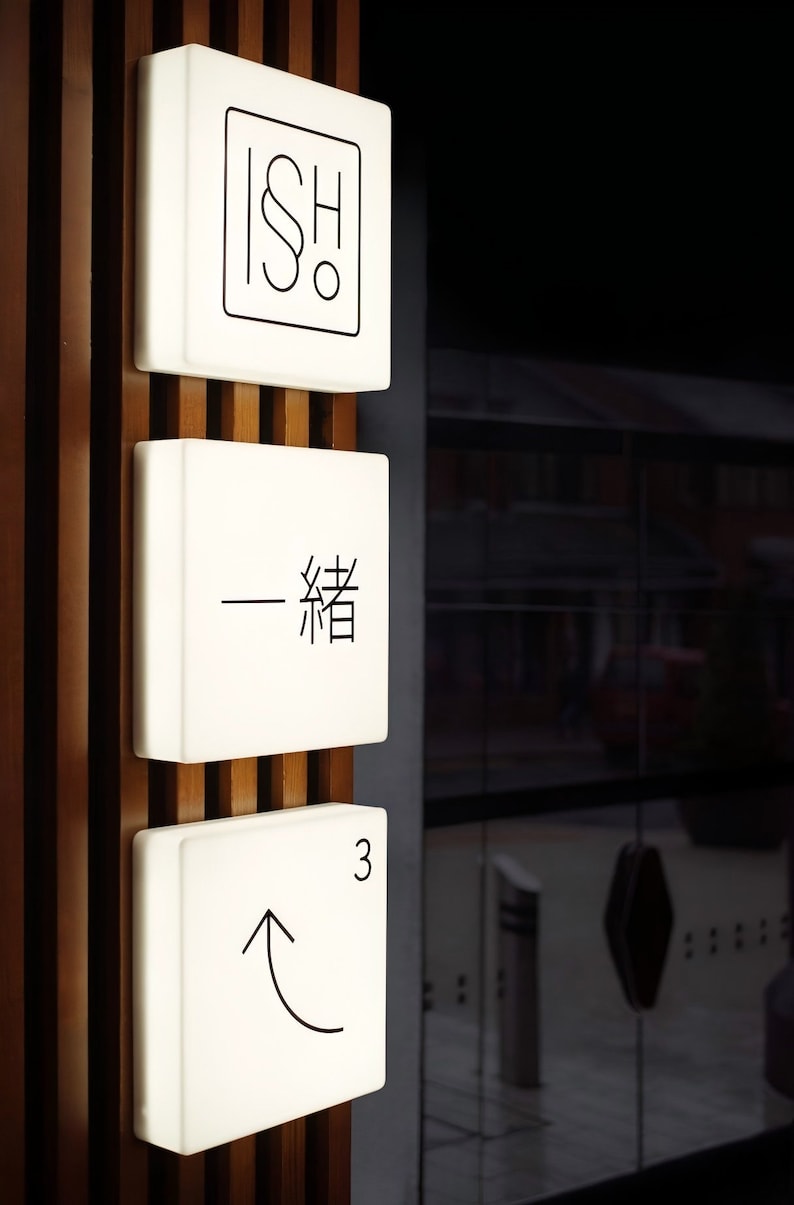 Op de afbeelding: Drie verlichte witte vierkante borden met zwarte tekst en symbolen zijn op een houten muur gemonteerd. Het bovenste bord heeft de letters "ISH" met een symbool. Het middelste bord heeft een Chinees karakter. Het onderste bord heeft het nummer "3" en een pijl omhoog.