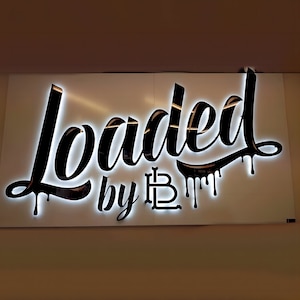 K&ouml;nnte beinhalten: Ein wei&szlig;es Schild mit schwarzer Beschriftung, auf dem in einer stilisierten Schrift "Loaded by LB" steht. Die Buchstaben sind wei&szlig; umrandet und haben einen tropfenden Effekt.