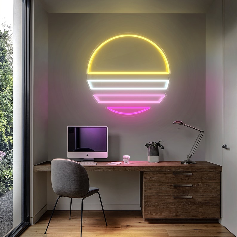 Op de afbeelding: Een modern thuiskantoor met een bureau en een computer. De muur achter het bureau heeft een neonlicht in de vorm van een zonsondergang met gele, witte en roze tinten.