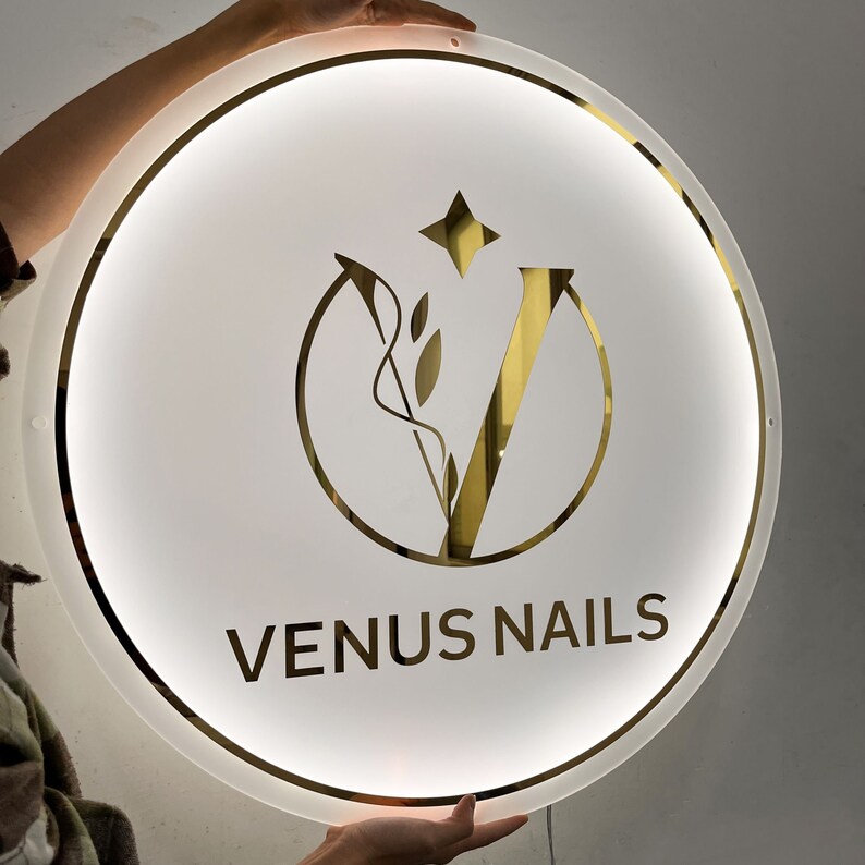 May include: A white round sign with a gold outline and a gold stylized letter V with a leaf design inside. The text "VENUS NAILS" is written in gold below the letter V.