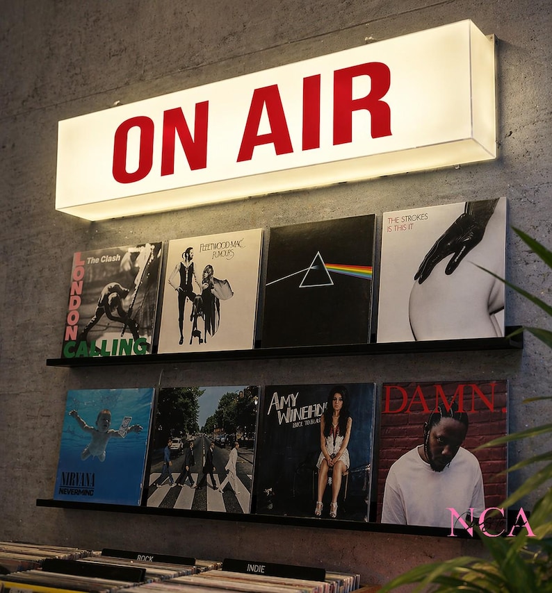 Op de afbeelding: Een rechthoekig, verlicht "ON AIR" bord met rode letters boven twee planken met vinylplaten. De albums zijn onder meer "London Calling" van The Clash, "Rumours" van Fleetwood Mac en "DAMN" van Kendrick Lamar.
