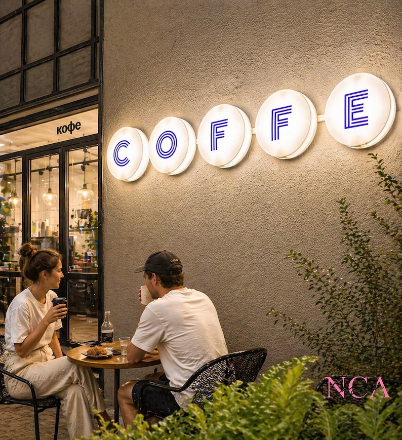 Puede incluir: Escena de un caf&eacute; al aire libre al anochecer. La palabra "COFFE" se muestra en letreros circulares iluminados con letras azules. Una pareja est&aacute; sentada en una mesa peque&ntilde;a, bebiendo bebidas. El caf&eacute; tiene grandes ventanales y un exterior de tonos neutros.