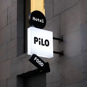 Puede incluir: Letrero exterior para un hotel y restaurante. El letrero est&aacute; montado en una pared de piedra y presenta un letrero circular negro que dice "Hotel" en blanco. Debajo hay un letrero rectangular blanco con el nombre "Pilo" en negro. Una flecha negra apunta a "FOOD".
