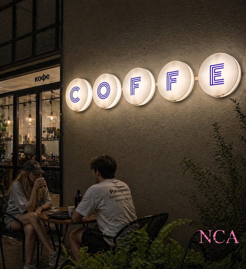 Op de afbeelding: Buitenkant van een caf&eacute; 's nachts. De letters "COFFEE" worden gevormd door verlichte, ronde borden met blauwe letters. Een stel zit aan een tafel en geniet van drankjes. Het caf&eacute; heeft grote ramen en een deur.