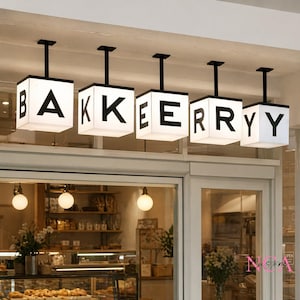 Op de afbeelding: Vijf verlichte, vierkante lichtarmaturen met de tekst "BAKERYY" hangen boven een bakkerij-ingang. Elke witte kubus heeft een zwarte letter. De gevel van de bakkerij is hieronder zichtbaar, met glazen deuren en vitrines vol bakkerijproducten.
