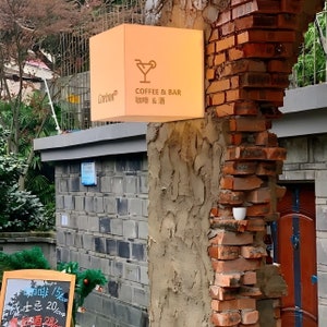 Op de afbeelding: Een vierkant, verlicht bord voor een koffie- en bar. Het bord is beige en toont een cocktailglas-icoon, de woorden "COFFEE & BAR" en Chinese karakters. Het bord is gemonteerd op een bakstenen en betonnen structuur. Het woord "Carbon" staat er ook op.