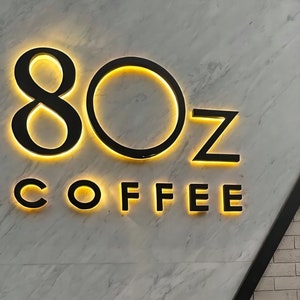 K&ouml;nnte beinhalten: Ein schwarzes und gelbes Schild mit dem Text "8OZ COFFEE" auf einem wei&szlig;en Marmorhintergrund.