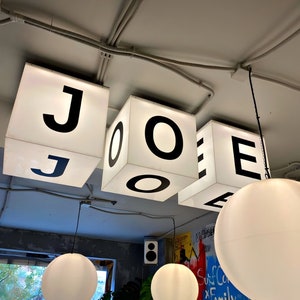 Puede incluir: Interior de un caf&eacute; con letreros c&uacute;bicos iluminados que deletrean "JOE". Debajo, cuelgan l&aacute;mparas colgantes de globo blanco, que complementan la est&eacute;tica brillante y moderna. El fondo incluye una ventana y arte mural colorido.
