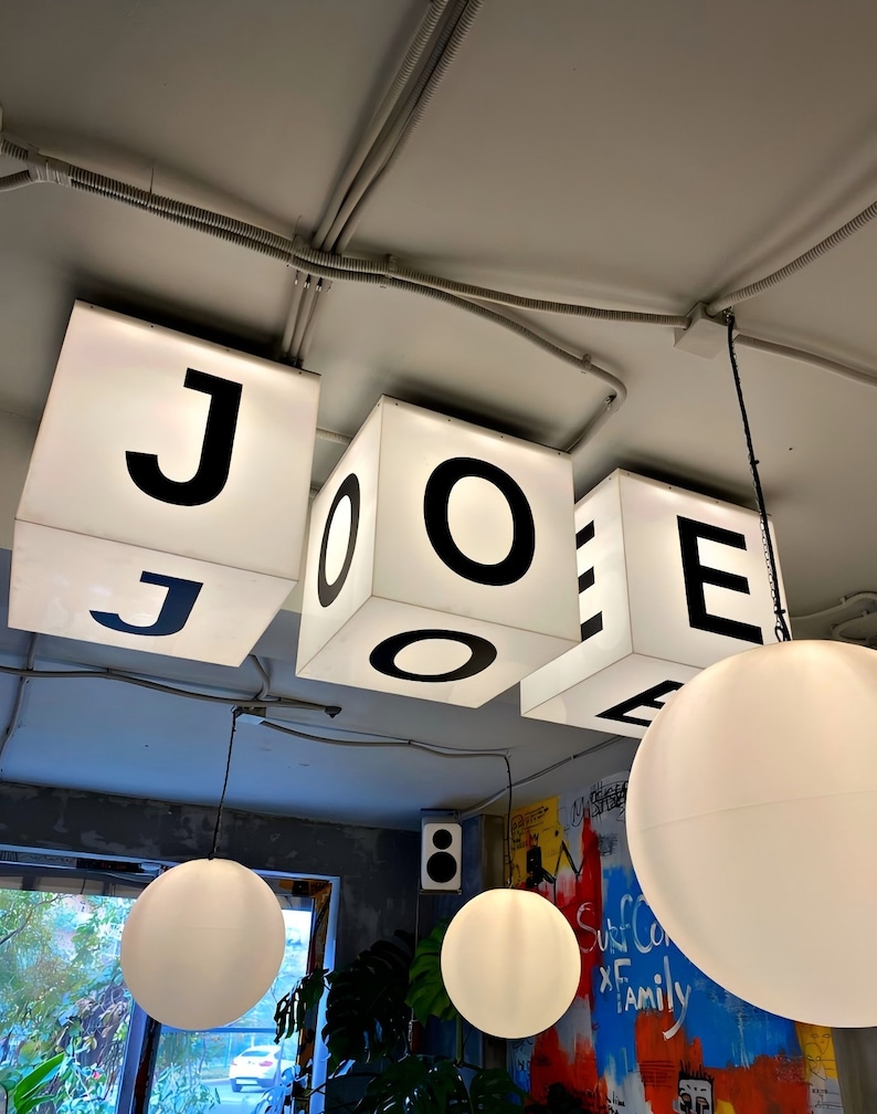 Puede incluir: Interior de un caf&eacute; con letreros c&uacute;bicos iluminados que deletrean "JOE". Debajo, cuelgan l&aacute;mparas colgantes de globo blanco, que complementan la est&eacute;tica brillante y moderna. El fondo incluye una ventana y arte mural colorido.