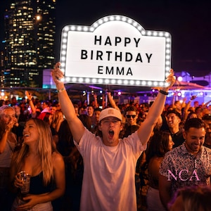 Puede incluir: Un letrero luminoso rectangular estilo marquesina con fondo blanco y letras negras que dicen HAPPY BIRTHDAY EMMA. El letrero cuenta con un borde de pequeñas bombillas LED de color blanco brillante y se sostiene en alto en una multitud nocturna.