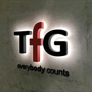 K&ouml;nnte beinhalten: Ein wei&szlig;es Schild mit schwarzen Buchstaben, die "TFG" buchstabieren, und dem Satz "everybody counts" in schwarzen Buchstaben darunter. Das "F" ist rot.
