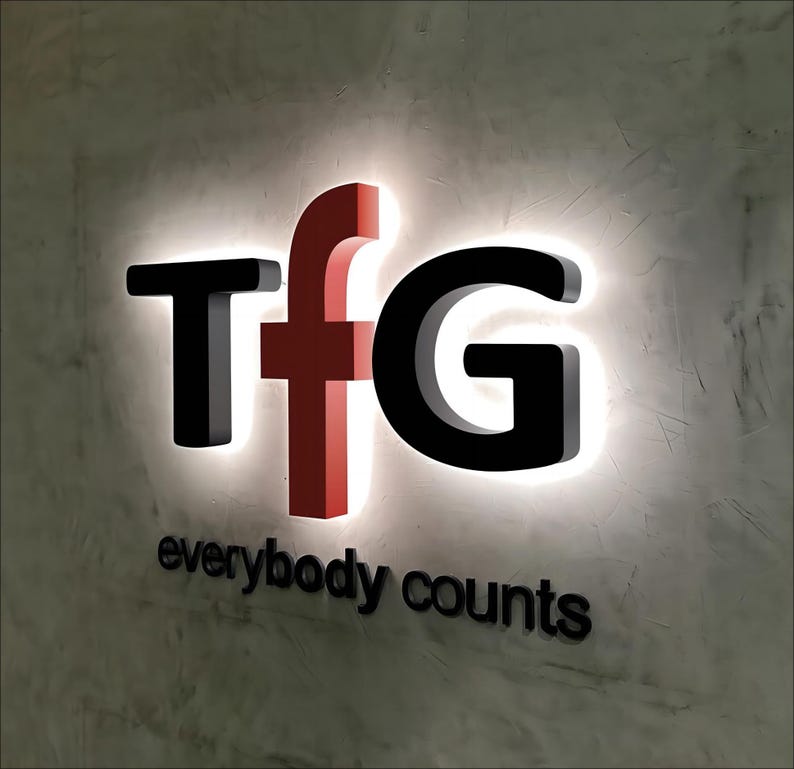 K&ouml;nnte beinhalten: Ein wei&szlig;es Schild mit schwarzen Buchstaben, die "TFG" buchstabieren, und dem Satz "everybody counts" in schwarzen Buchstaben darunter. Das "F" ist rot.