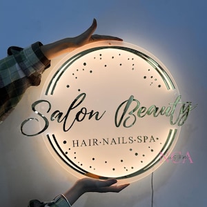 May include: A round, white, illuminated sign with a gold border and gold lettering that reads "Salon Beauty" with the text "HAIR.NAILS.SPA" underneath.