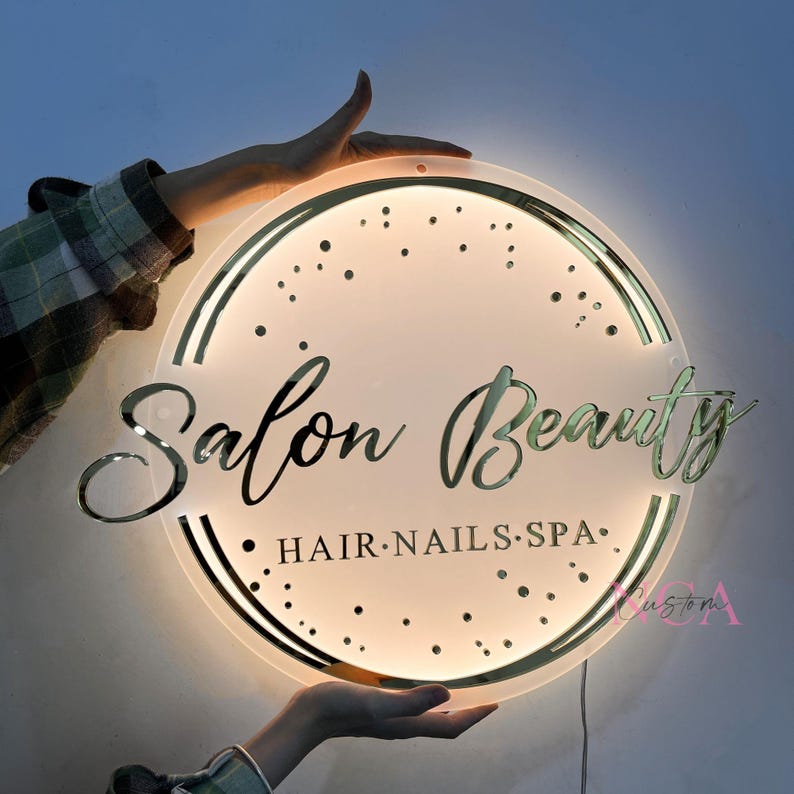 May include: A round, white, illuminated sign with a gold border and gold lettering that reads "Salon Beauty" with the text "HAIR.NAILS.SPA" underneath.