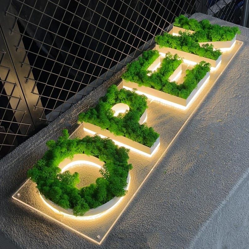 May include: A white sign with the word "OPEN" spelled out in green moss. The letters are illuminated with white lights.