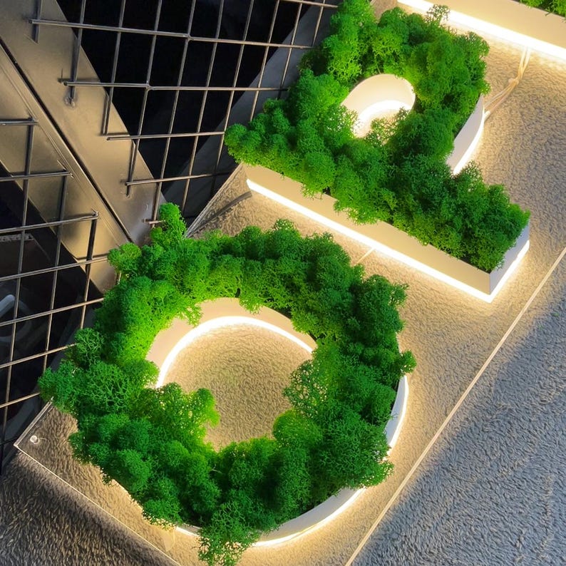 May include: A white sign with a green moss border, shaped like the letter "O". The sign is illuminated with a white light.