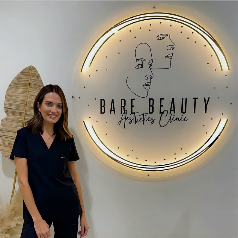 May include: A woman in a black scrub top stands in front of a white sign with a gold circle around it. The sign says "Bare Beauty Aesthetics Clinic" and has a line drawing of two faces.
