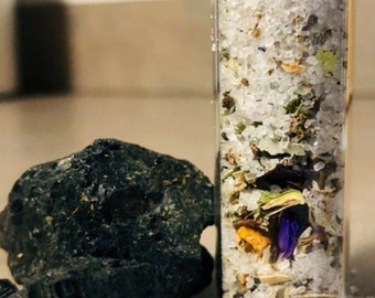May include: A glass jar filled with white bath salts, dried flowers, and herbs. The jar has a silver lid and is sitting on a white surface with black rocks.