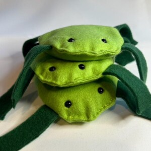 May include: A handmade plush toy in the shape of a stacked, segmented creature. The toy is primarily lime green with dark green appendages and black bead eyes. The plush is made of soft fabric and has a whimsical design.