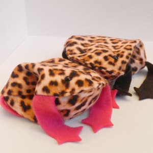May include: Two plush leopard print frog toys with pink felt arms and black felt legs. One frog is lying down with its arms outstretched, while the other frog is standing up with its arms bent.